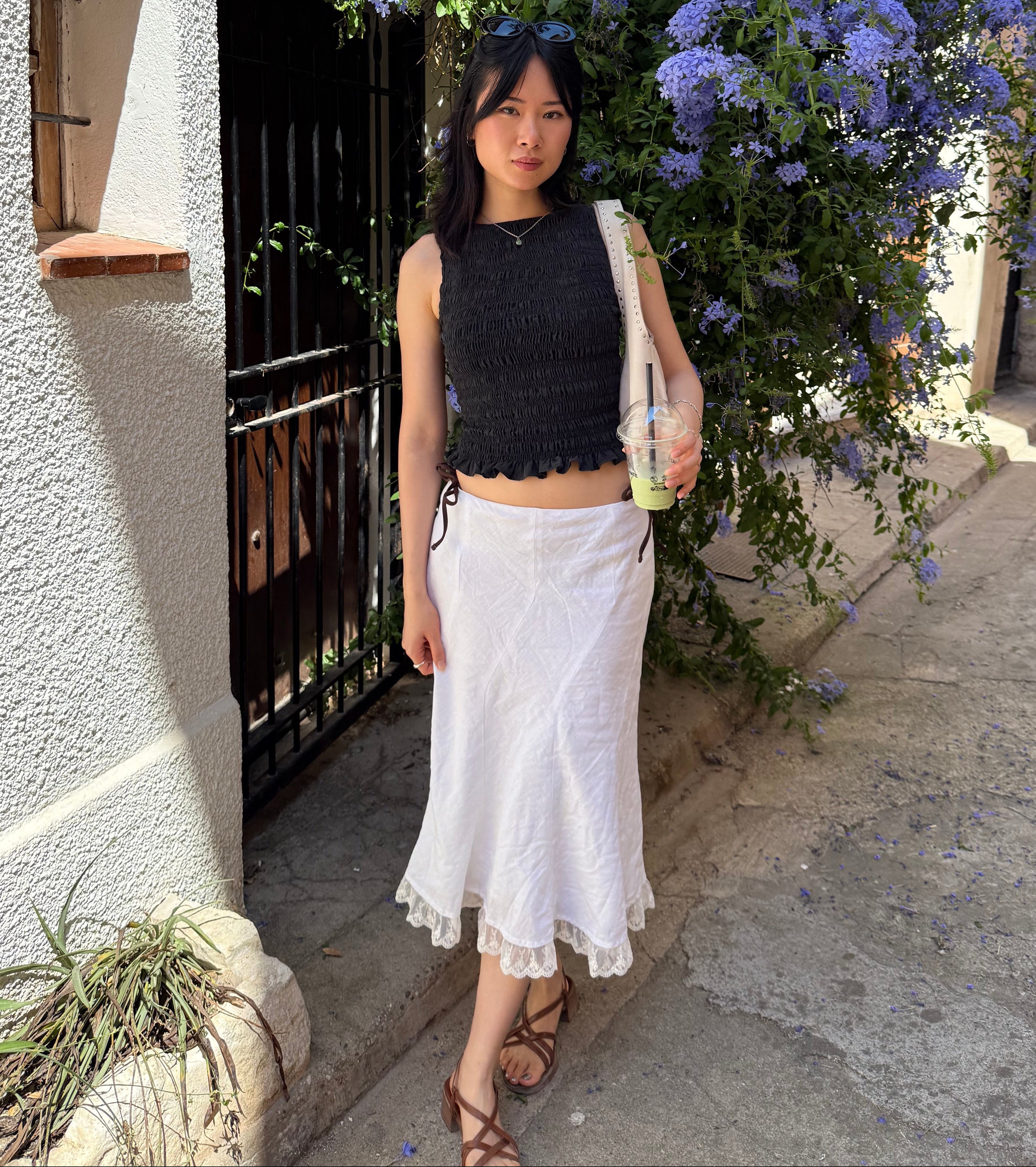 A woman stands outdoors in a sunlit alleyway wearing a sleeveless black cropped top and a white midi skirt, holding a drink in front of a large bush of blue flowers.