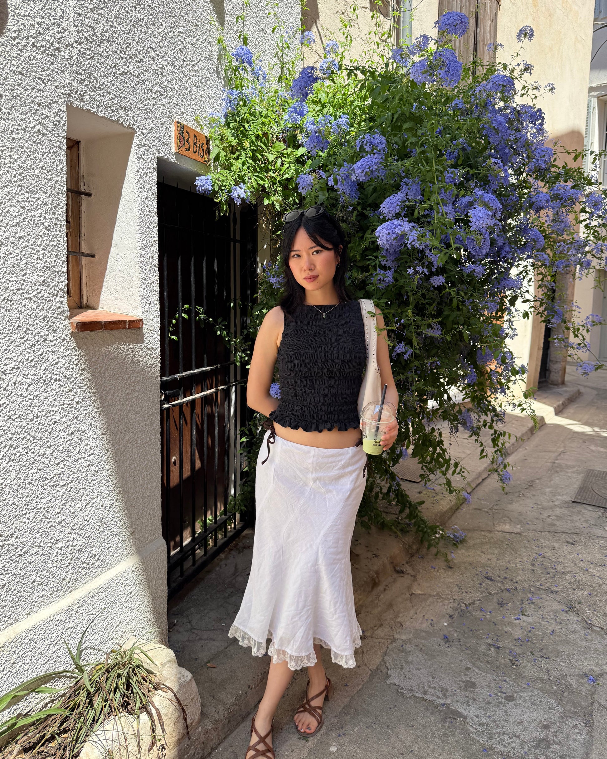 A woman posing outdoors in a black sleeveless cropped top and a white midi skirt, standing in front of a white stucco building with a large purple flower bush.