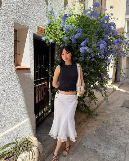 A woman posing outdoors in a black sleeveless cropped top and a white midi skirt, standing in front of a white stucco building with a large purple flower bush.