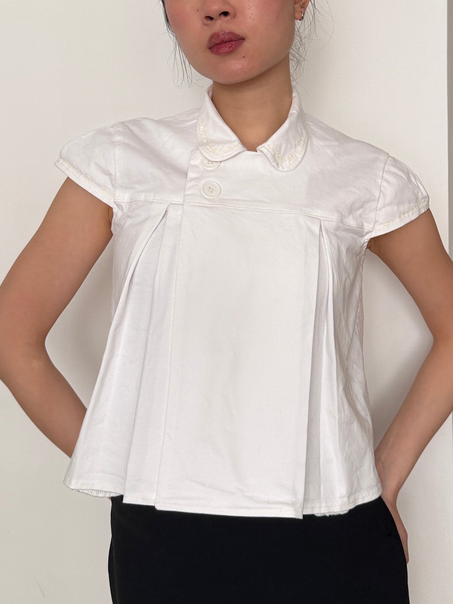 White short-sleeve blouse worn by a person on a plain background. Collar with side buttons.